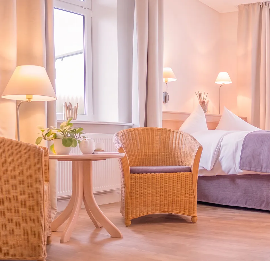 Bright hotel room corner with two wicker chairs around a round table, a plant and teapot, and a bed in the background.