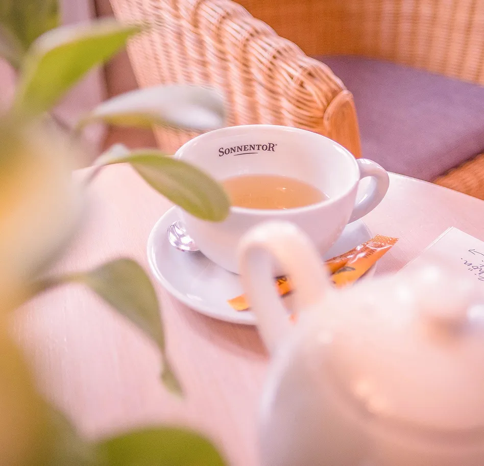 Cup of tea on a saucer with a Sonnentor label, on a light wooden table with a small packet nearby.
