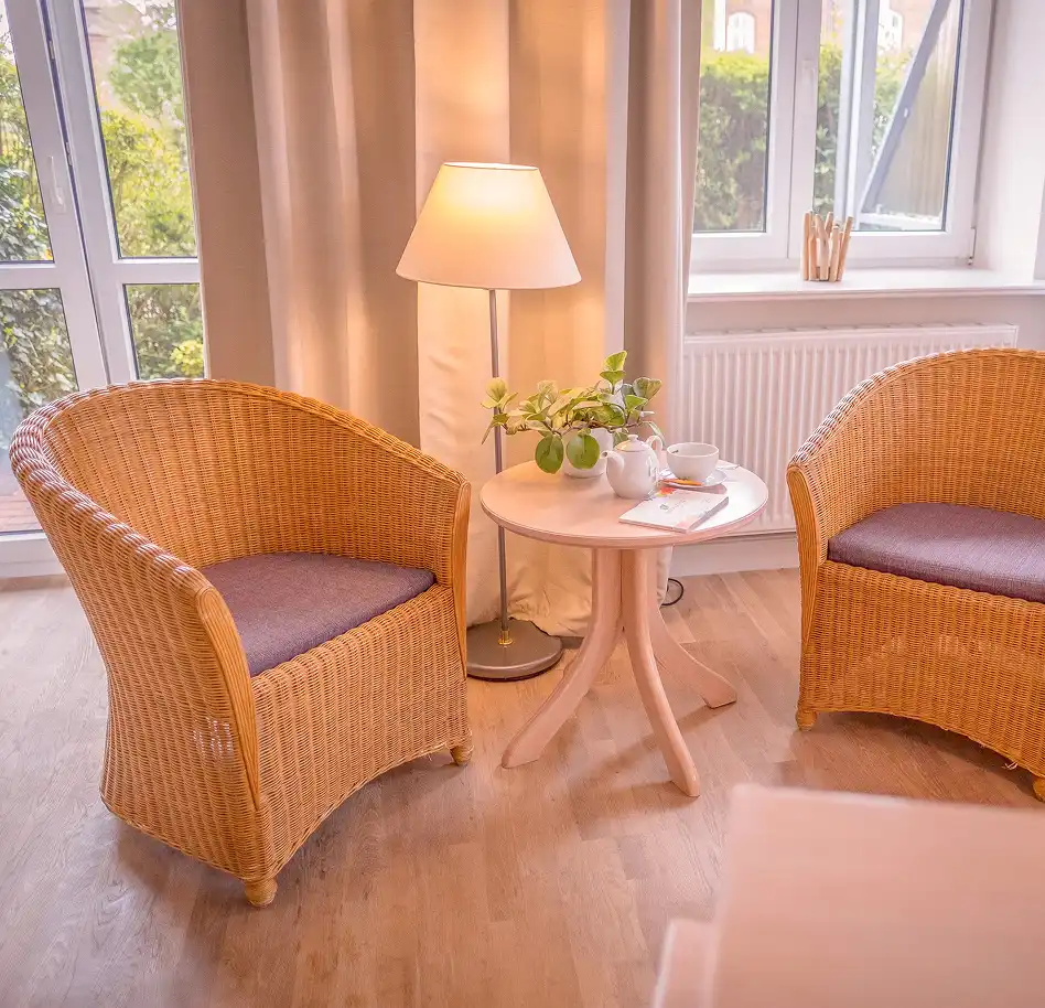 Cozy seating nook with two wicker armchairs, a round side table, a floor lamp, and a teacup setup by a windowed wall.