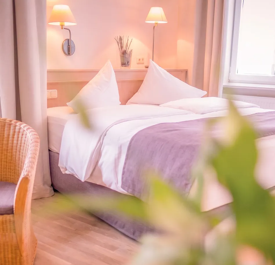 A neatly made bed with white sheets and lavender blanket in a softly lit hotel room, with two wall lamps and a wicker chair nearby.