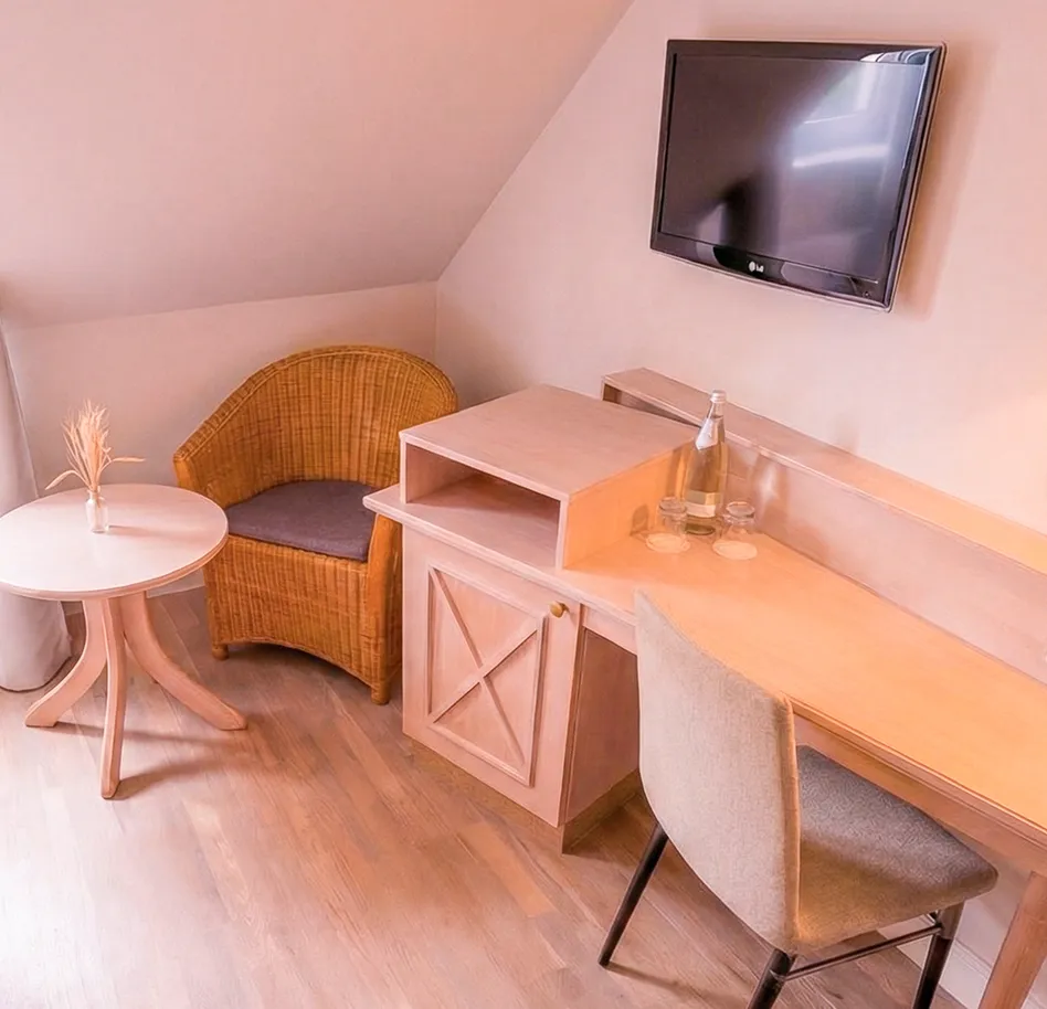 Wood-toned hotel room workspace: desk with chair, mounted TV, wicker armchair, and round side table with a vase.