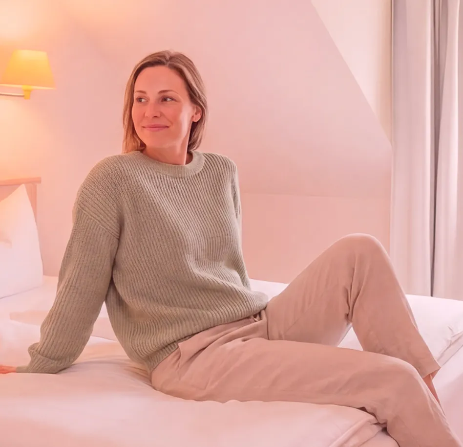Woman sits on a bed in a cozy beige knit sweater and light pants, looking to the left in a softly lit bedroom.