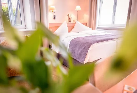 Bright hotel room with a neatly made bed, white linens, a purple throw, and warm bedside lamps by large windows.