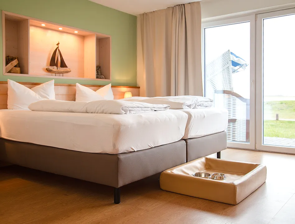 Hotel room with two beds, a beige pet bed and bowls on the floor, and a balcony door letting in light.