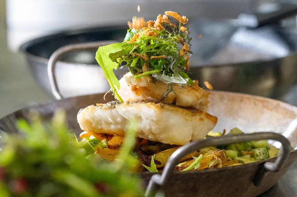 Three stacked fried white fish fillets topped with greens and crispy onions, resting on sautéed vegetables in a skillet.
