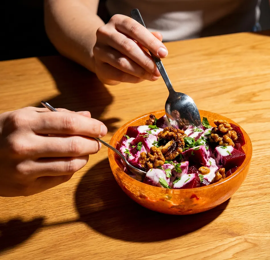 A person using a spoon and fork to eat a beet salad with yogurt dressing and walnuts from a wooden bowl on a table.