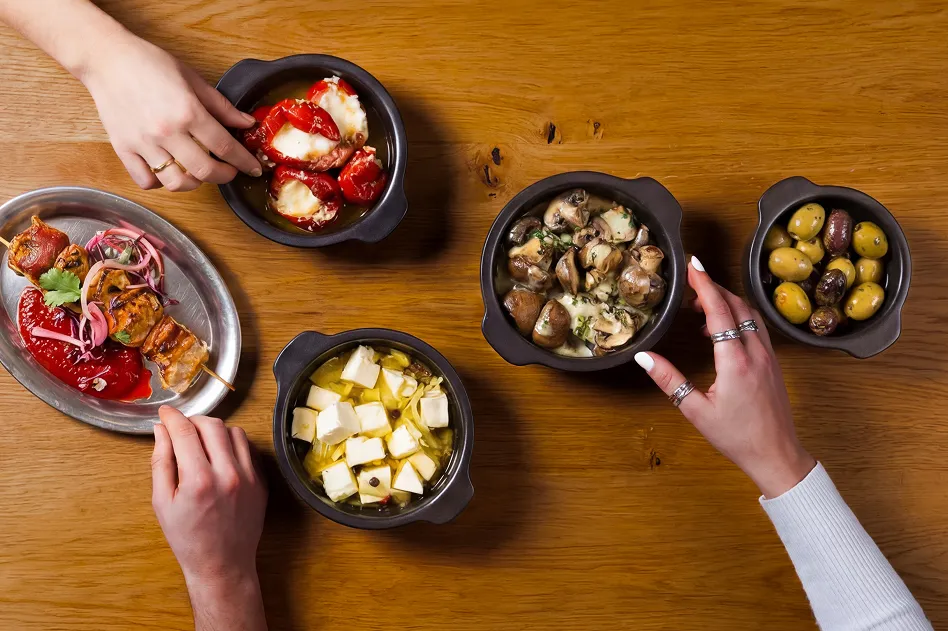 Assorted tapas bowls on a wooden table, with olives, mushrooms, feta cheese, roasted peppers with cheese, and skewered veggies; hands reach in.