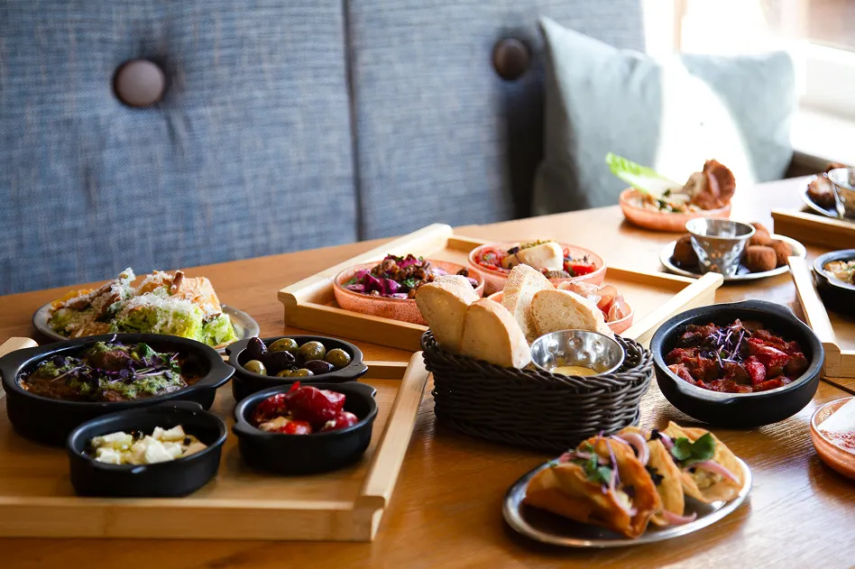 Table spread of assorted tapas: small bowls, olives, and bread baskets on a wooden table in a cozy restaurant setting
